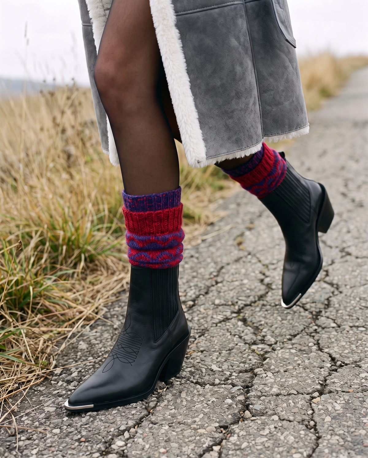 ASADLIE Nella black leather western ankle boots with metal toe tips, worn with red and indigo knitted wool socks and a grey suede shearling coat on a highland road