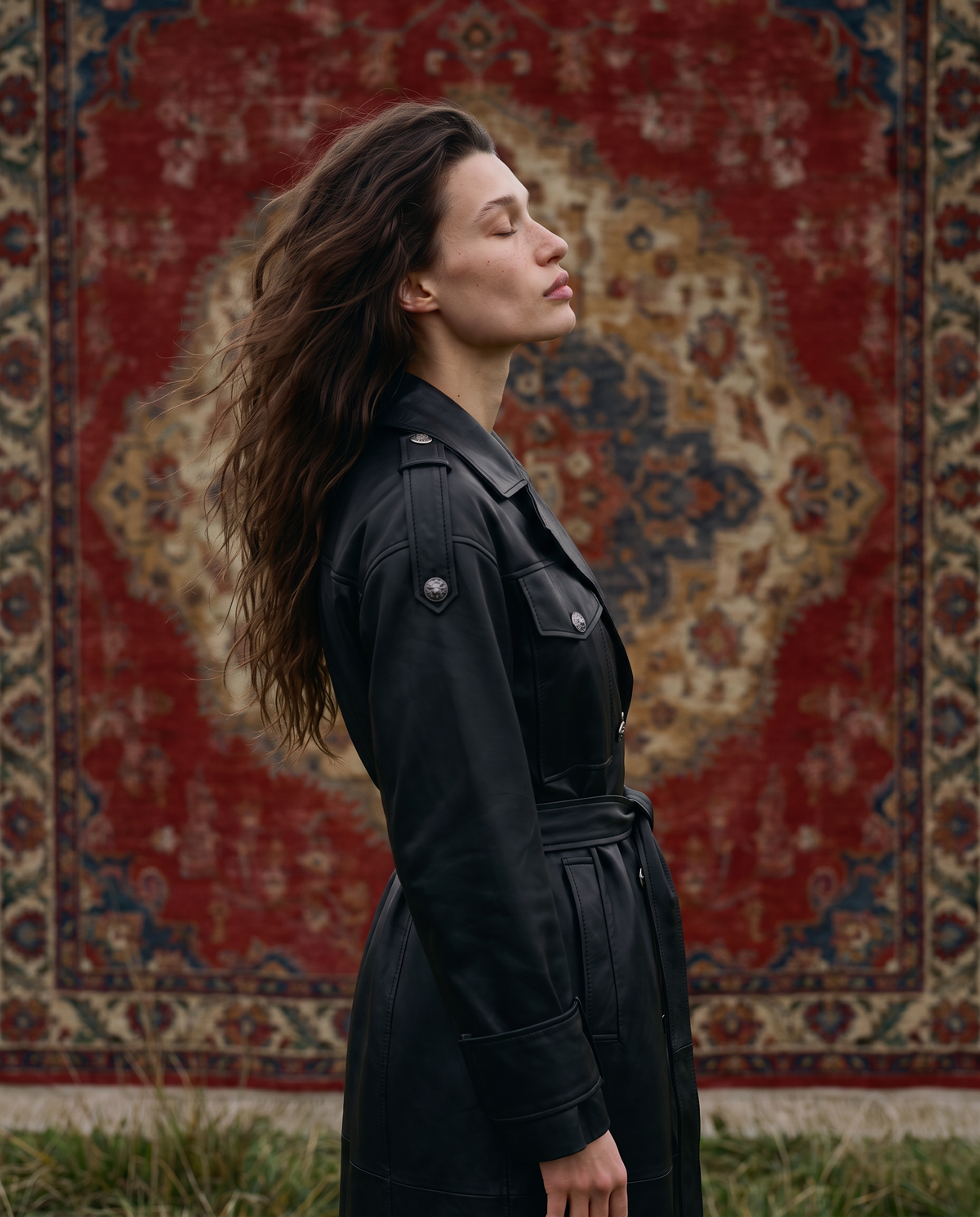 Woman in the ASADLIE Victoria long black leather coat in side profile, wind in her hair, in front of an old Azerbaijani carpet in the Guba highlands